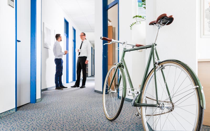 Bürobild mit Fahrrad im Vordergrund und Kollegen im Hintergrund